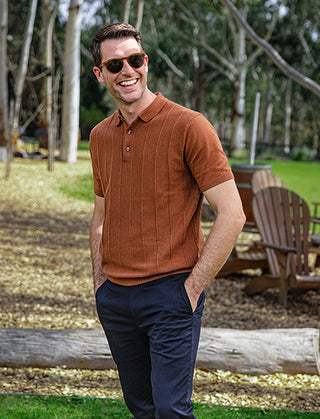 A man smiling wearing a brown polo top and navy chinos in a park